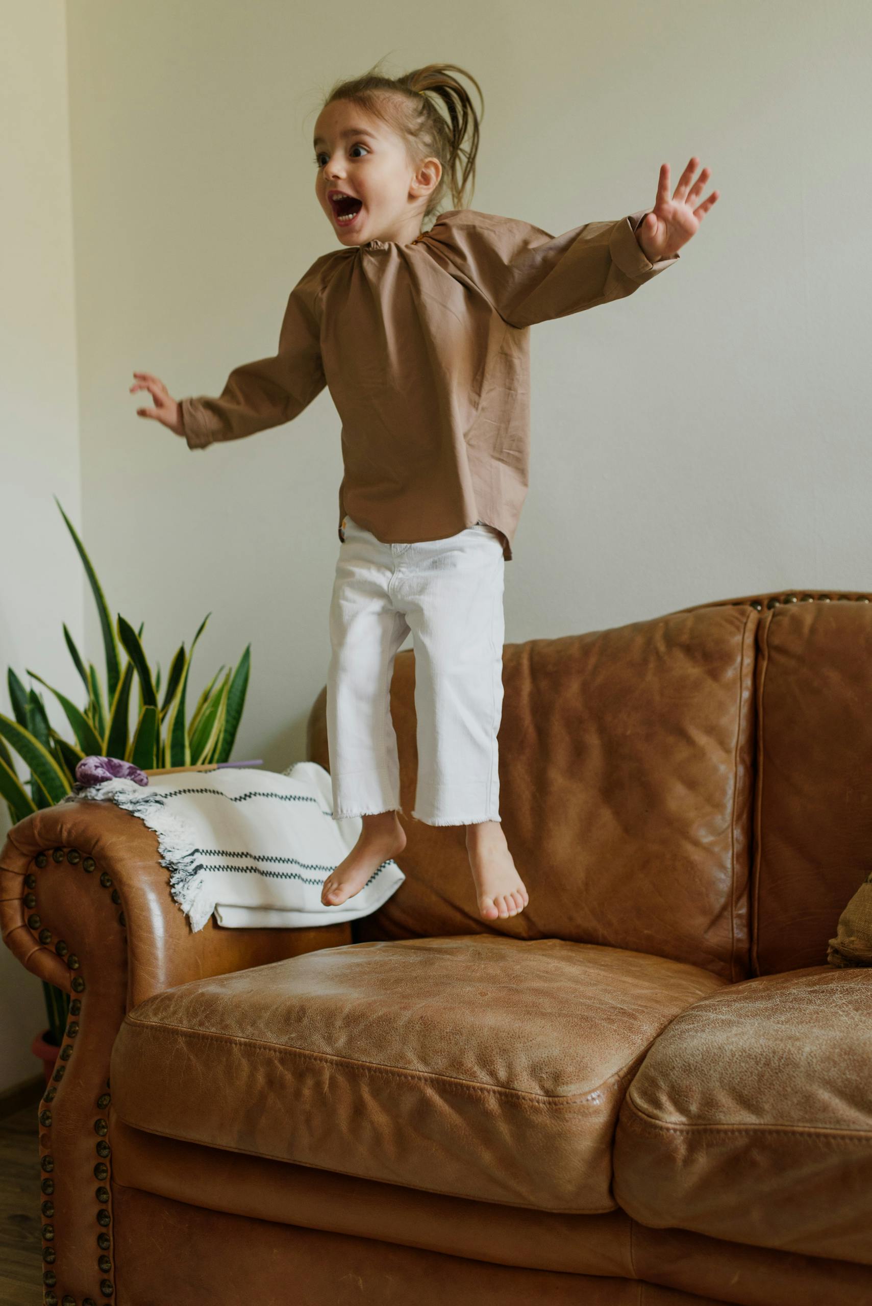 Happy child energetically jumping on a comfortable leather sofa indoors.